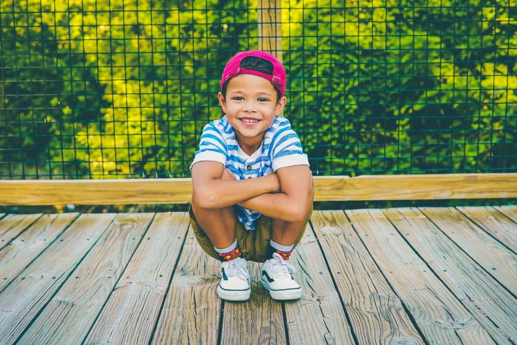 Brunette boy in a red hat and a blue and white striped shirt smiles as she crouches on a wooden pier