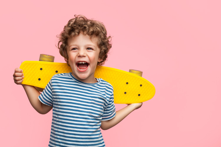 Curly-haired little boy holds a yellow skateboard and smiles against a pink wall