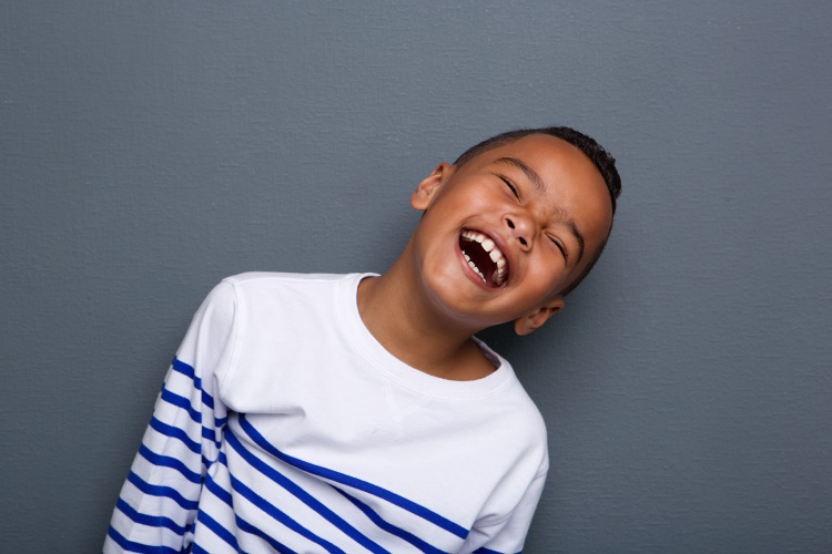 Dark-haired boy in a white and navy striped shirt smiles against a gray wall after visiting a children's dentist