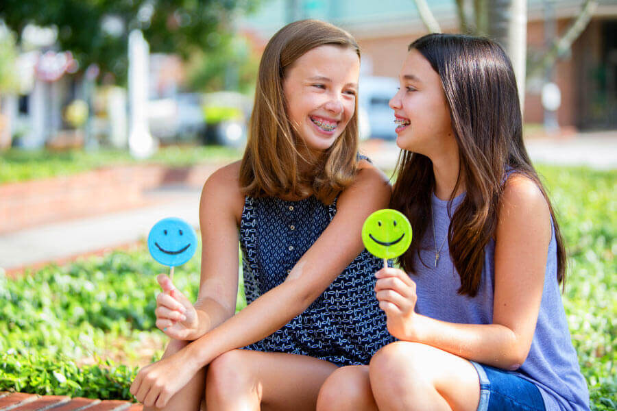 Two girls wear braces and smile at each other while sitting down with sugar-free lollipops in Tyler, TX