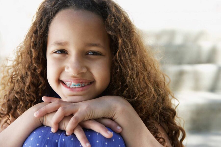 Brunette girl with interceptive orthodontics smiles with her hands on her knees outside her pediatric dentist in Tyler, TX