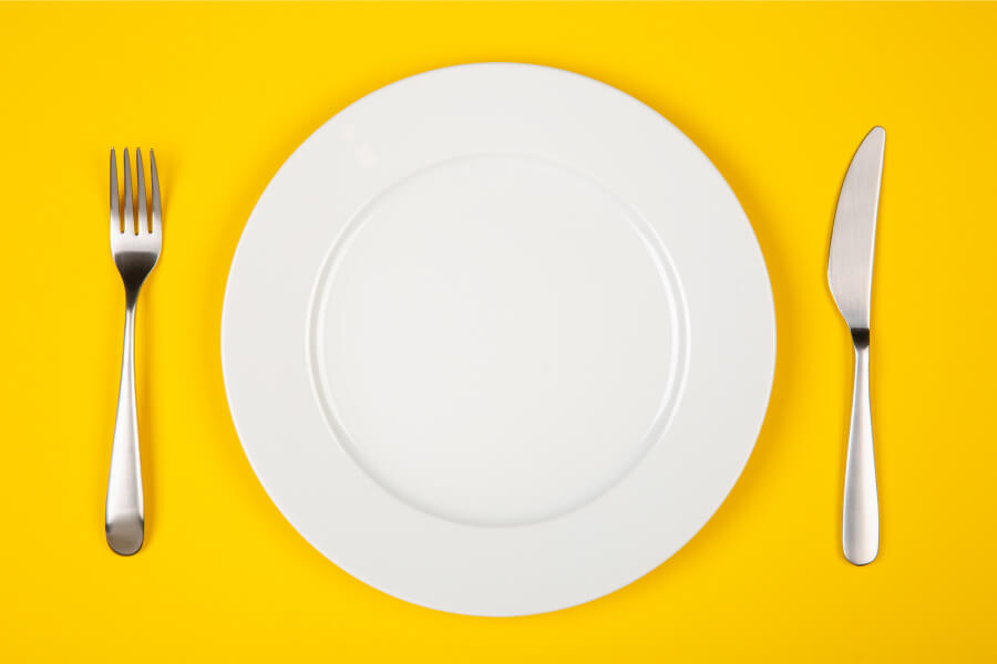 Aerial view of a white plate next to silverware on a yellow table waiting for Thanksgiving foods