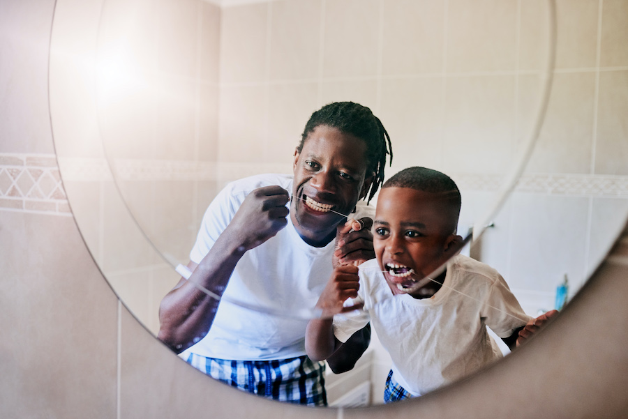 Black dad and son brush and floss their teeth while in their pajamas looking in the bathroom mirror