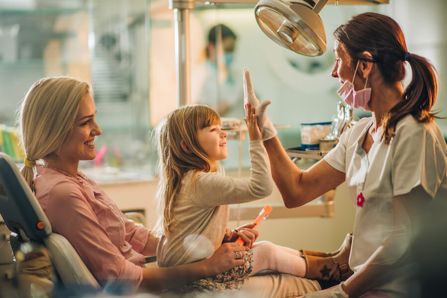 Brunette girl sitting on her mom's lap at the dentist gives her female hygienist a high five