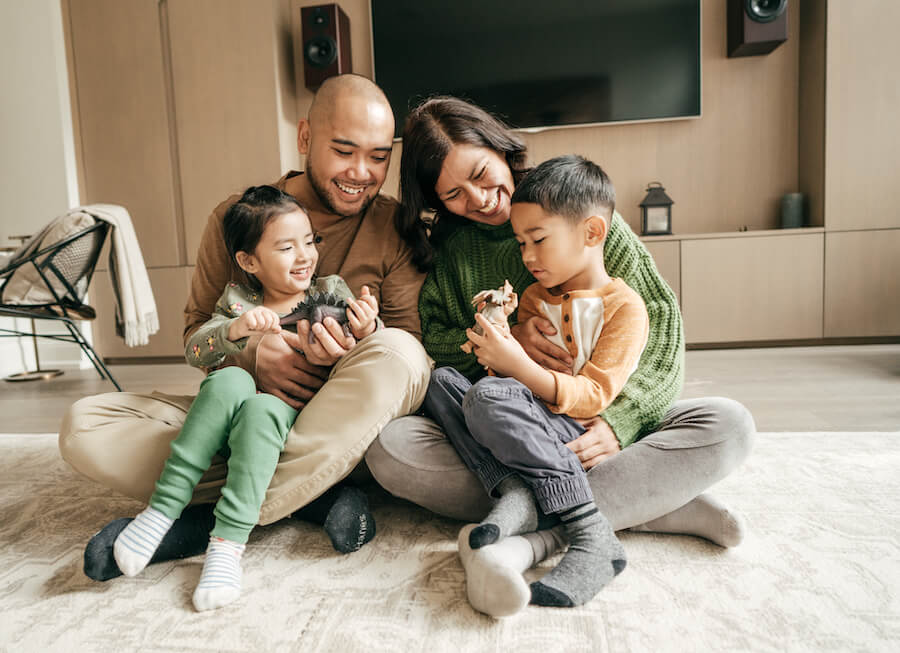 A mom and dad sit in their living room playing dinosaurs with their young son and daughter