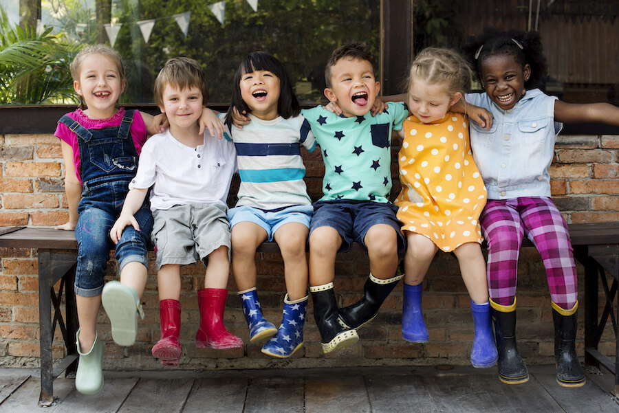 A group of 6 children smile as they sit together on a bench outside while wearing rainboots