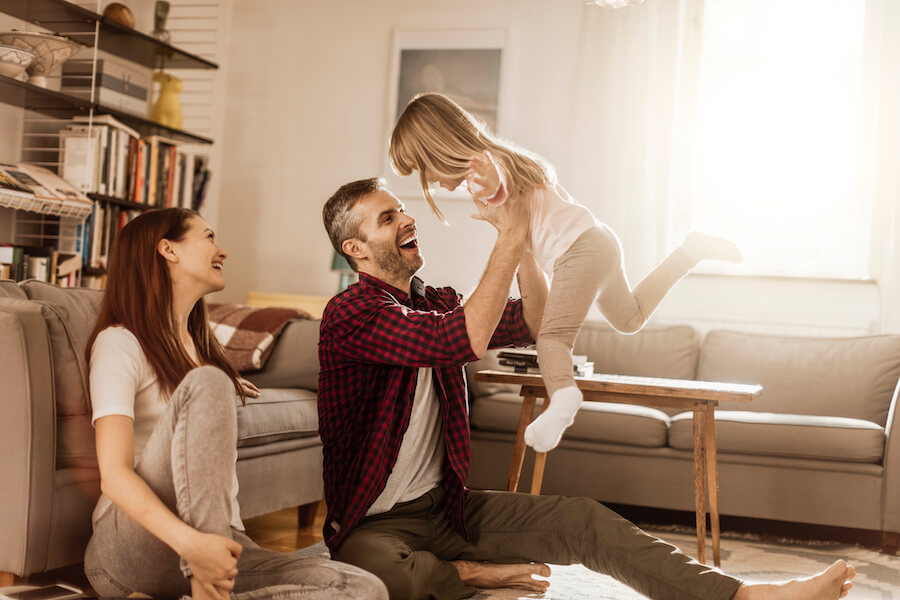 A mom smiles as dad picks up their young daughter as they play in their family room