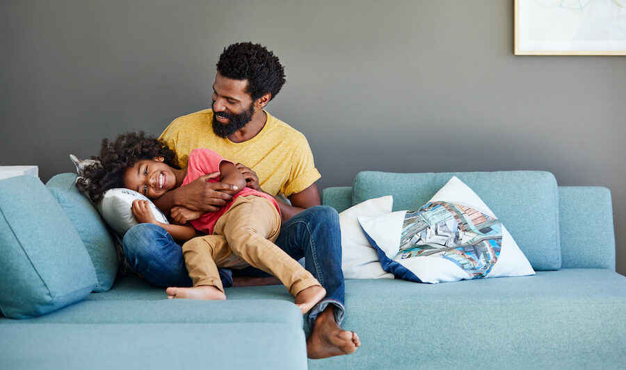 A Black dad in a yellow shirt smiles while playing with his son on the couch