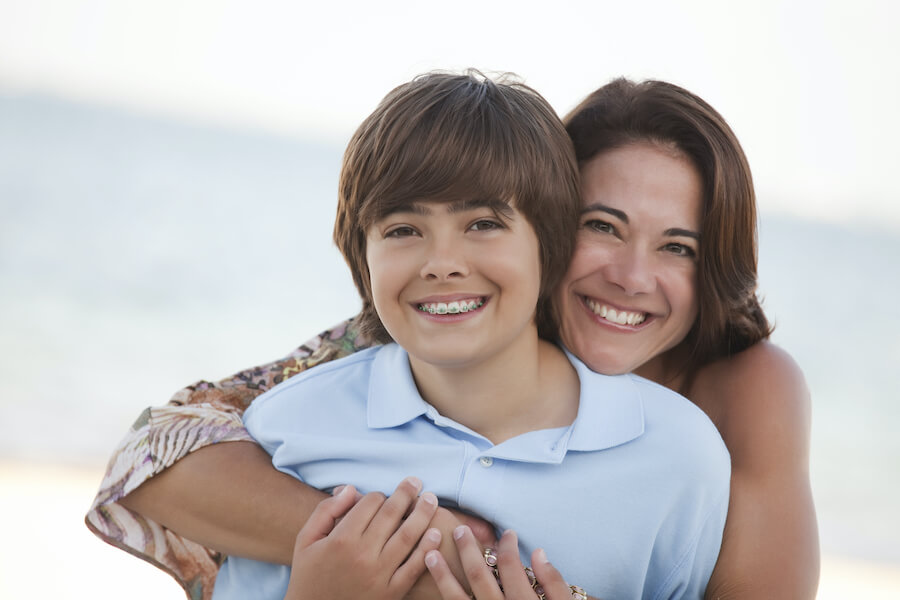 Brunette mom smiles while hugging her smiling brunette son with braces from behind