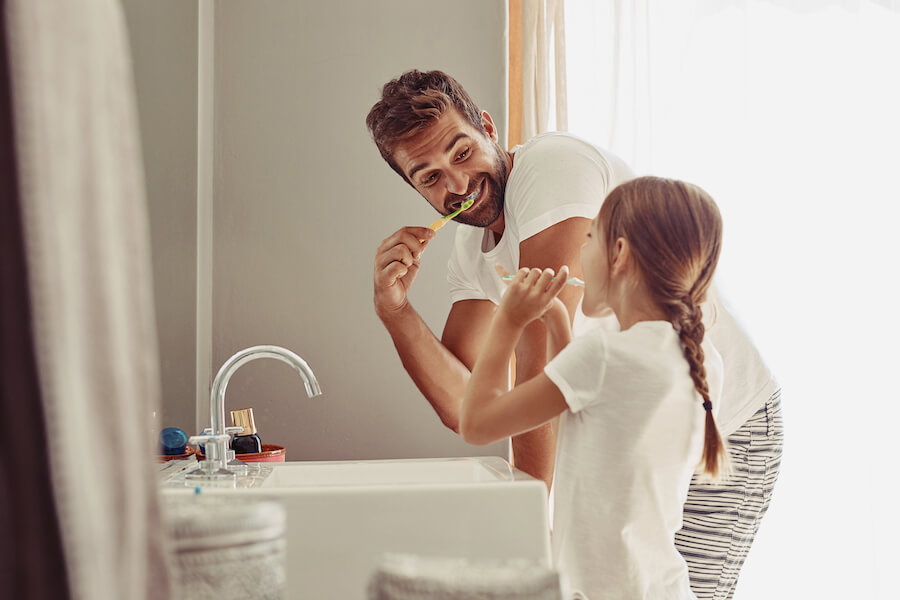 Photo of a smiling father brushing his teeth next to his daughter brushing her teeth, modeling the dental care tips.